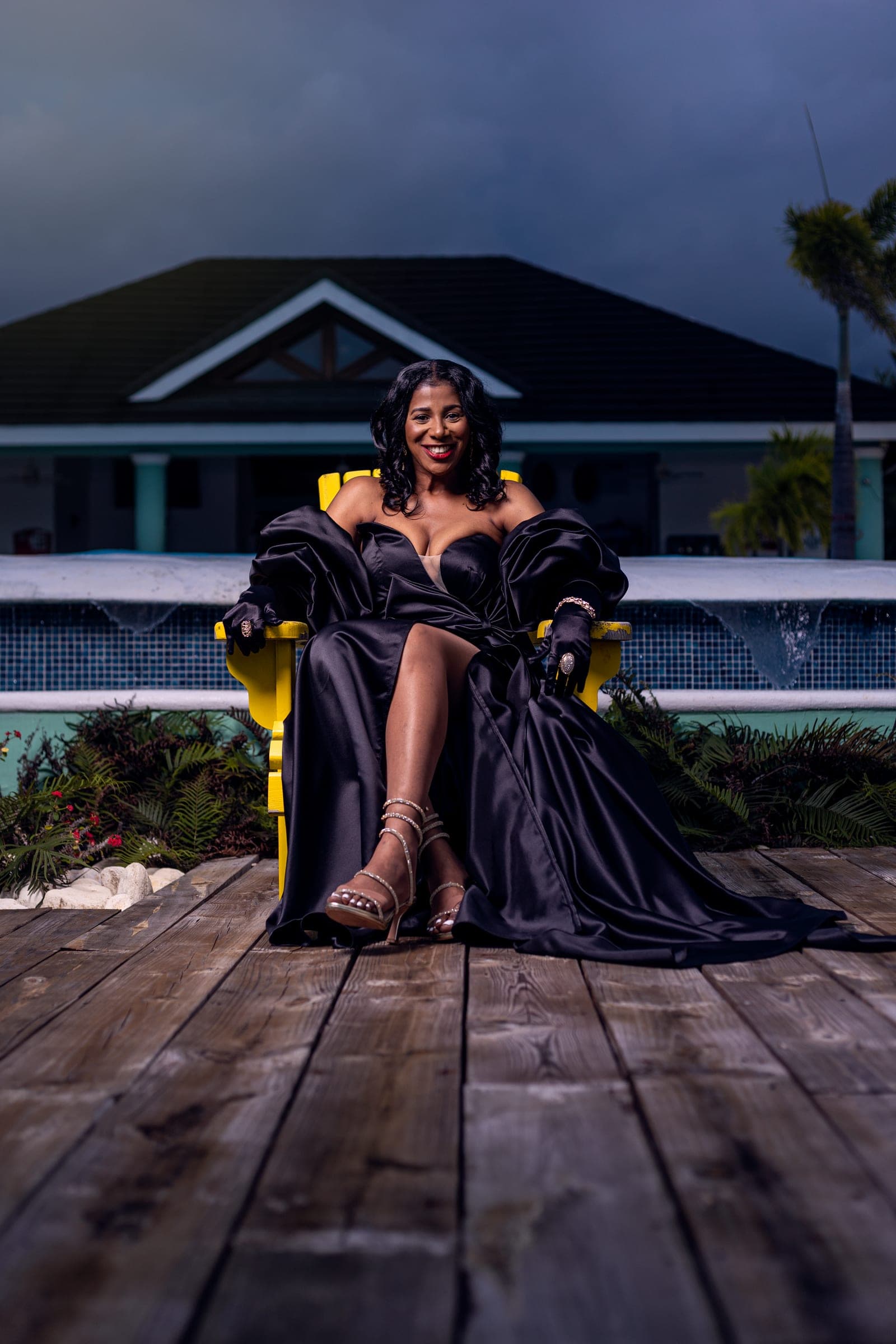 Woman in a black gown seated on a yellow chair by a pool at dusk